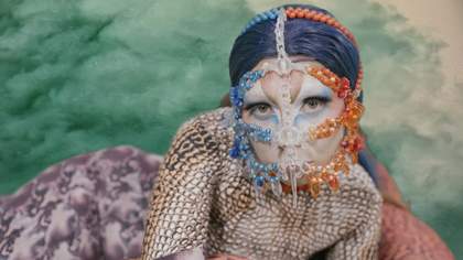Blue background. A woman lying down on her side. She is wearing a beaded head and face covering