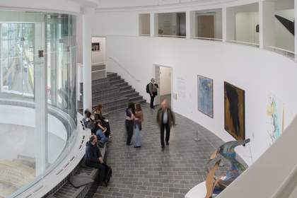 A curved room in the gallery with high ceilings, tall windows and large artworks hanging on the walls. One sculpture is large and stands on a plinth.